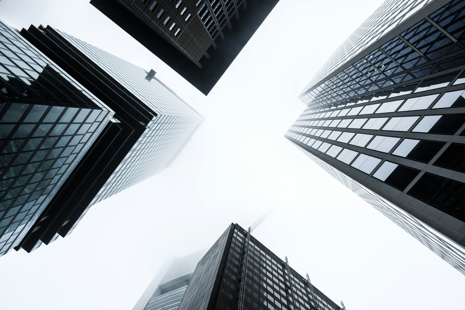 Low-angle view of glass and steel skyscrapers rising into a bright, misty sky.
